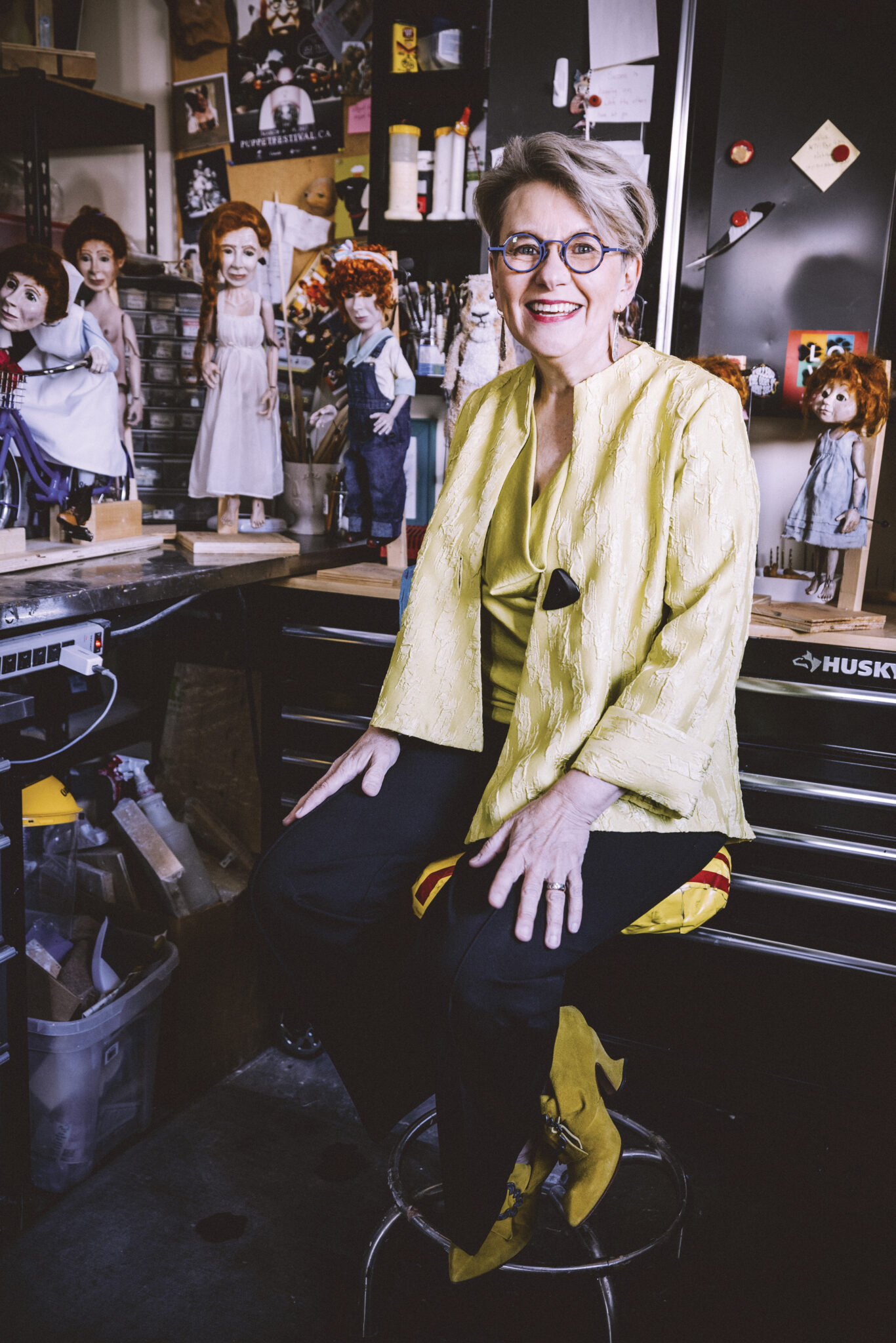 Juanita Dawn sits on a stool surrounded by puppets on a table.