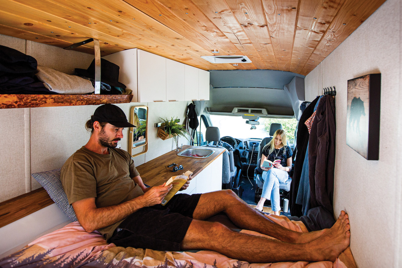 Two people sit adjacent to each other inside a camper van while they both read.