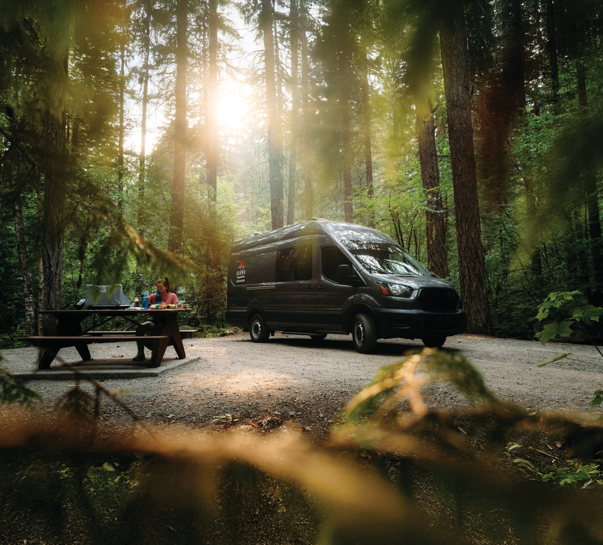 A woman sits at a picnic bench in front of a van in the forest.