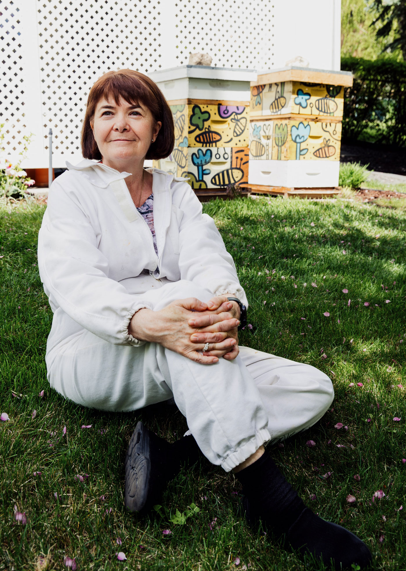 A woman sits on the ground in a beekeeping outfit with hives in the background.