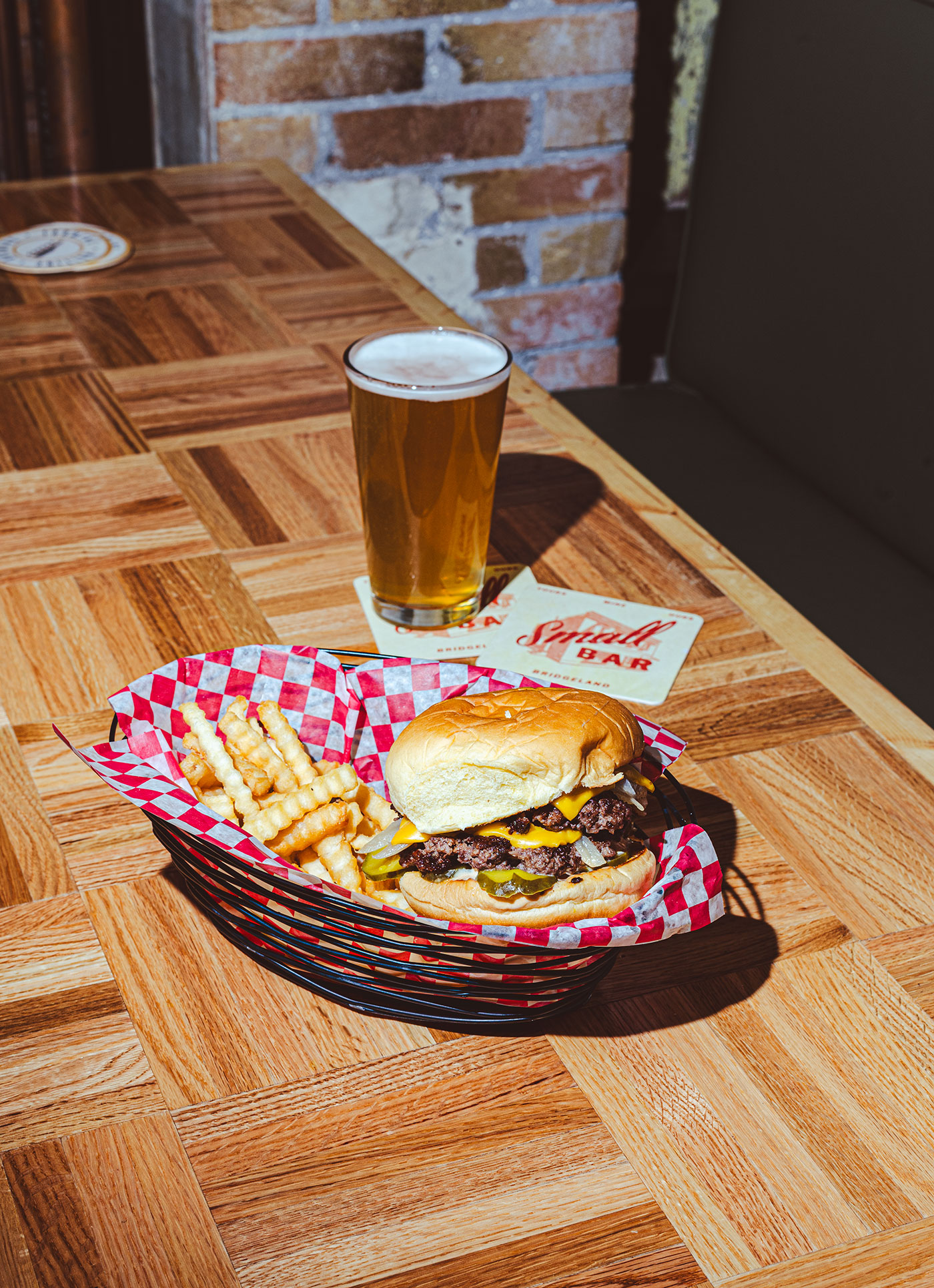 A basket with a cheeseburger and fries in the foreground and a glass of beer in the background.