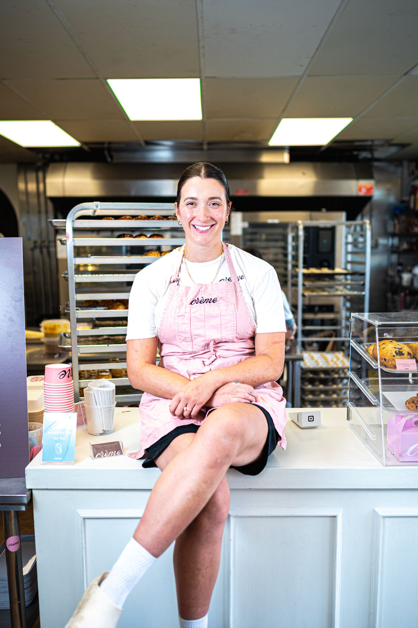 A chef in a pink apron sits on the counter in a bakery.