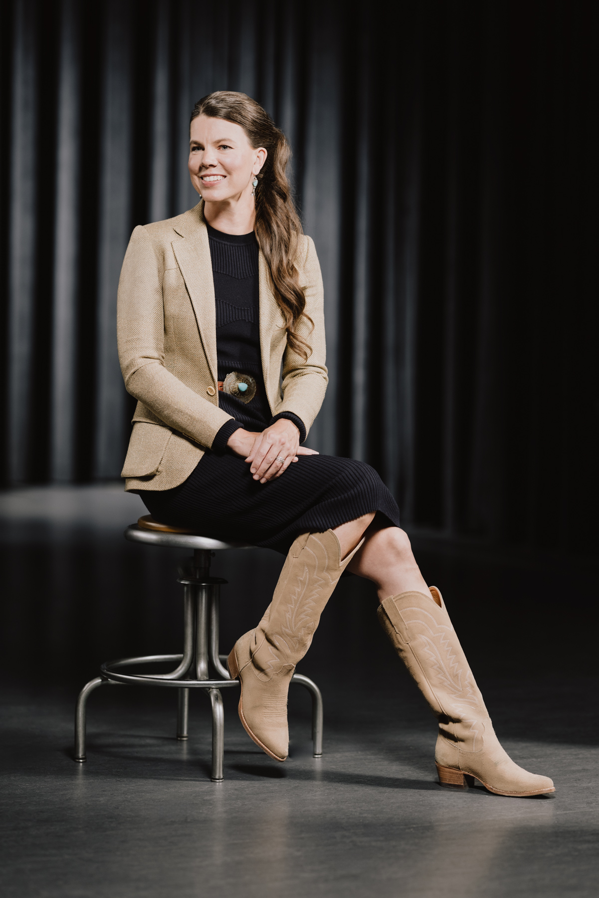 Kandis Twa sits wearing a tan blazer, a black belted dress and tall, pointed tan boots.
