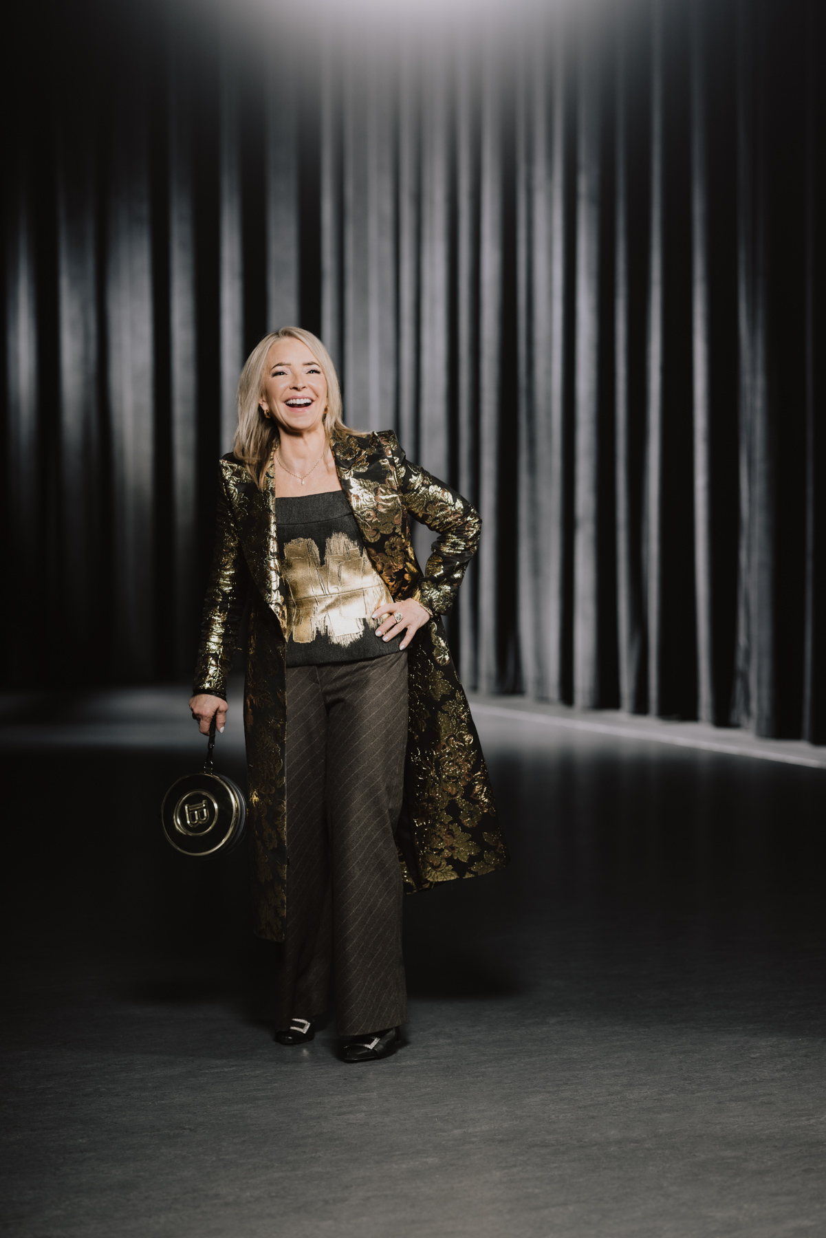 Cynthia P. Moore wearing a long gold-patterned jacket with a black shirt and gold graphic, black pants, boots and a circular black purse.