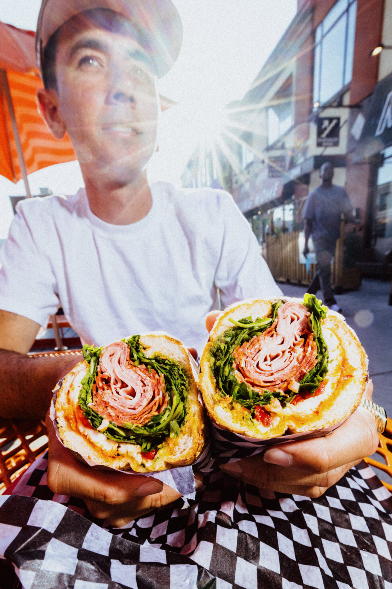 A person holds a sandwich in each hand as the sun shines behind him.