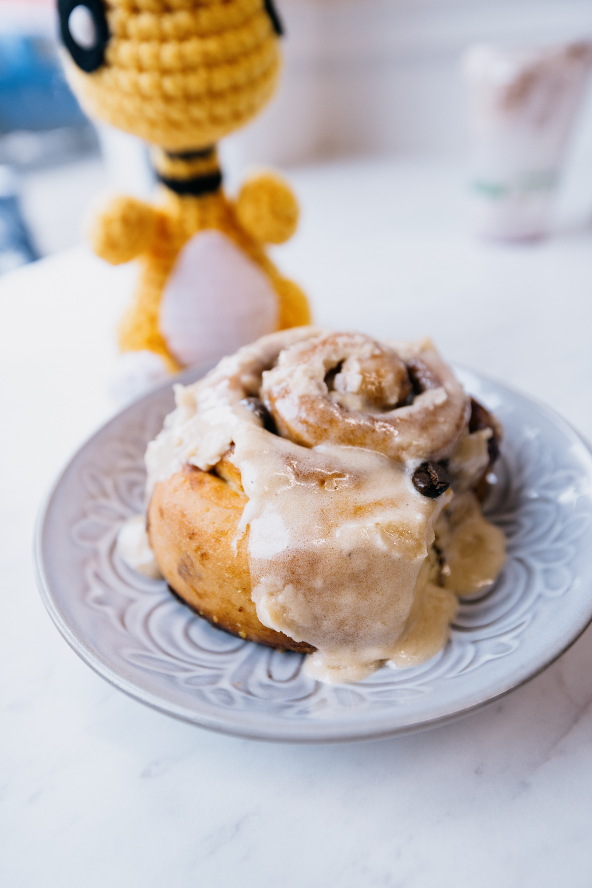 A cinnamon bun sits on top of a plate, with a crochet animal in the background.