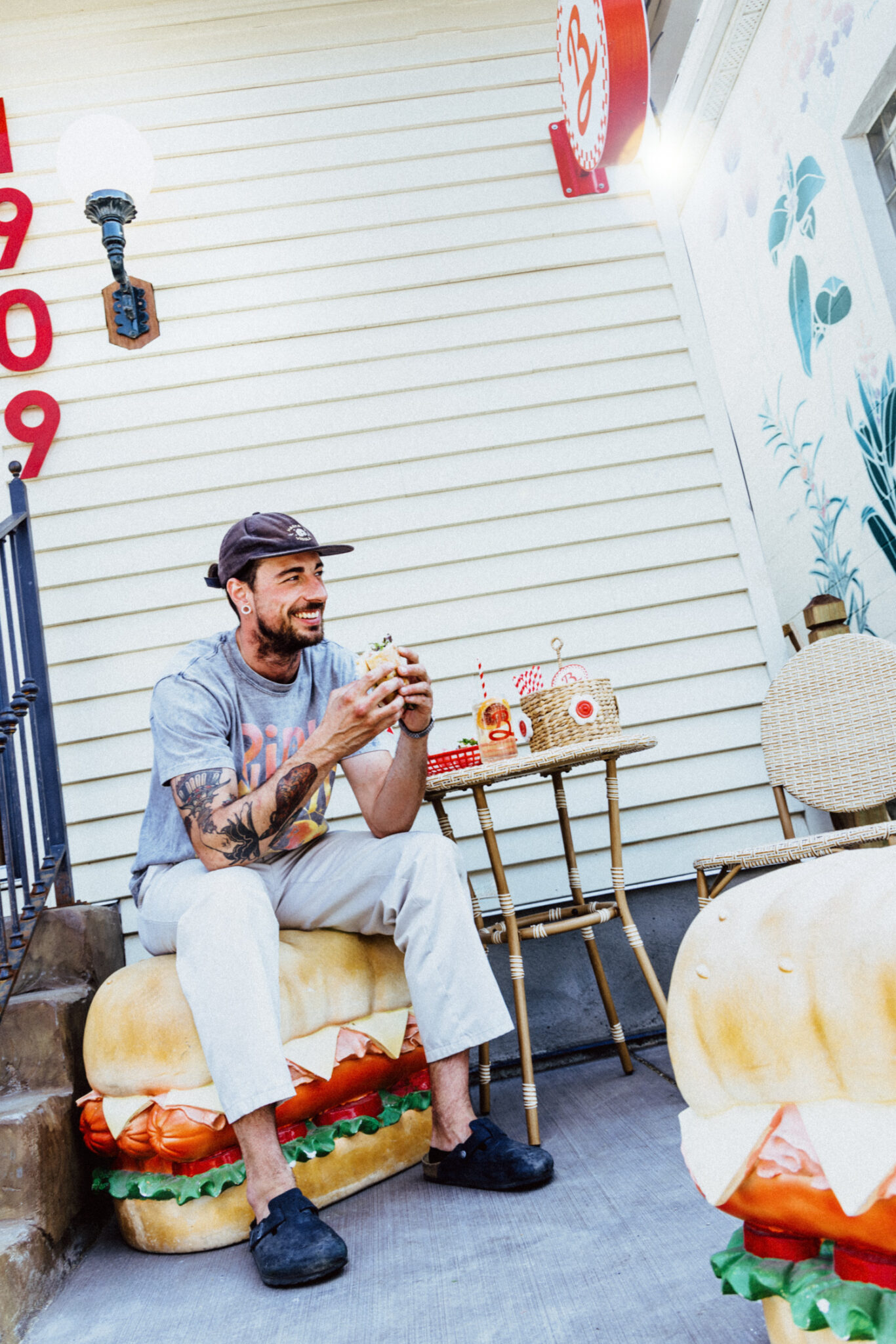 A man sits outside on a large bench that looks like a sandwich and eats a sandwich.