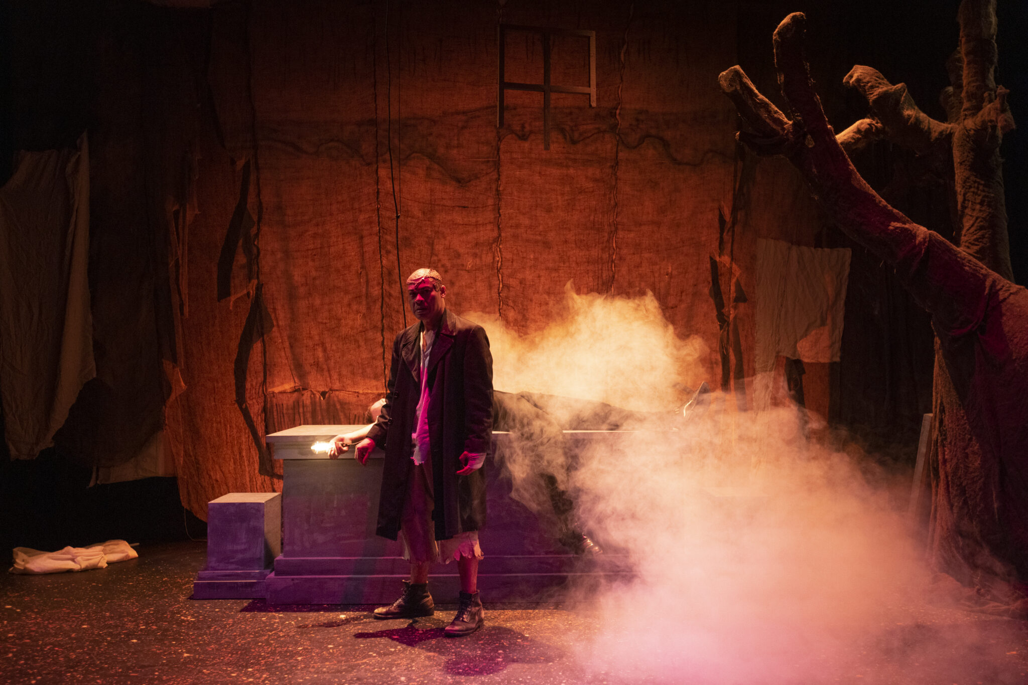 A performer surrounded by in red light and smoke on stage at a theatre production.