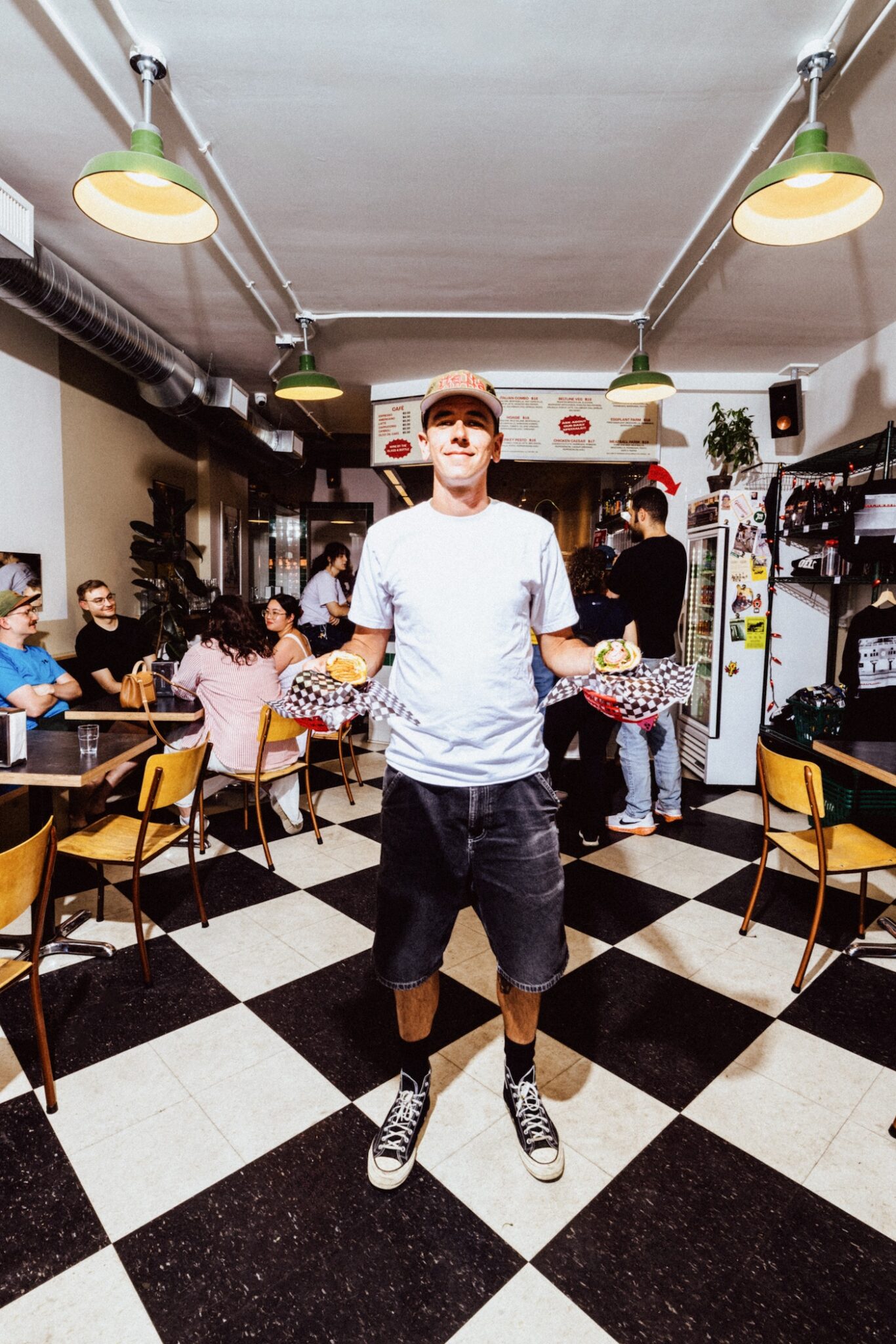 A person stands inside a sandwich shop holding a basket filled with a sandwich in each hand.