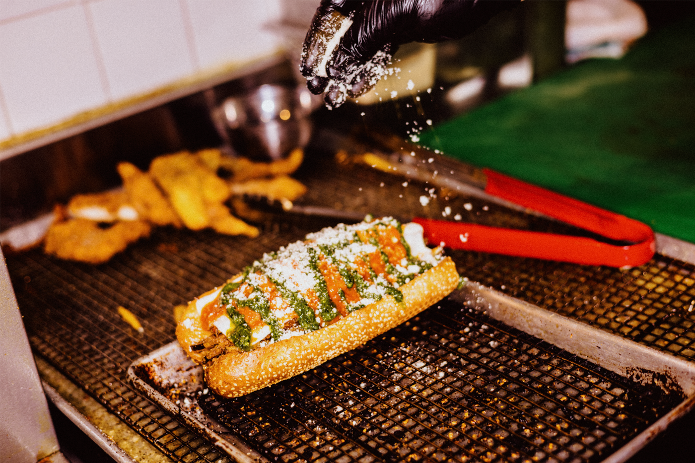 A gloved hand sprinkles cheese on top of a sandwich sitting on a tray.