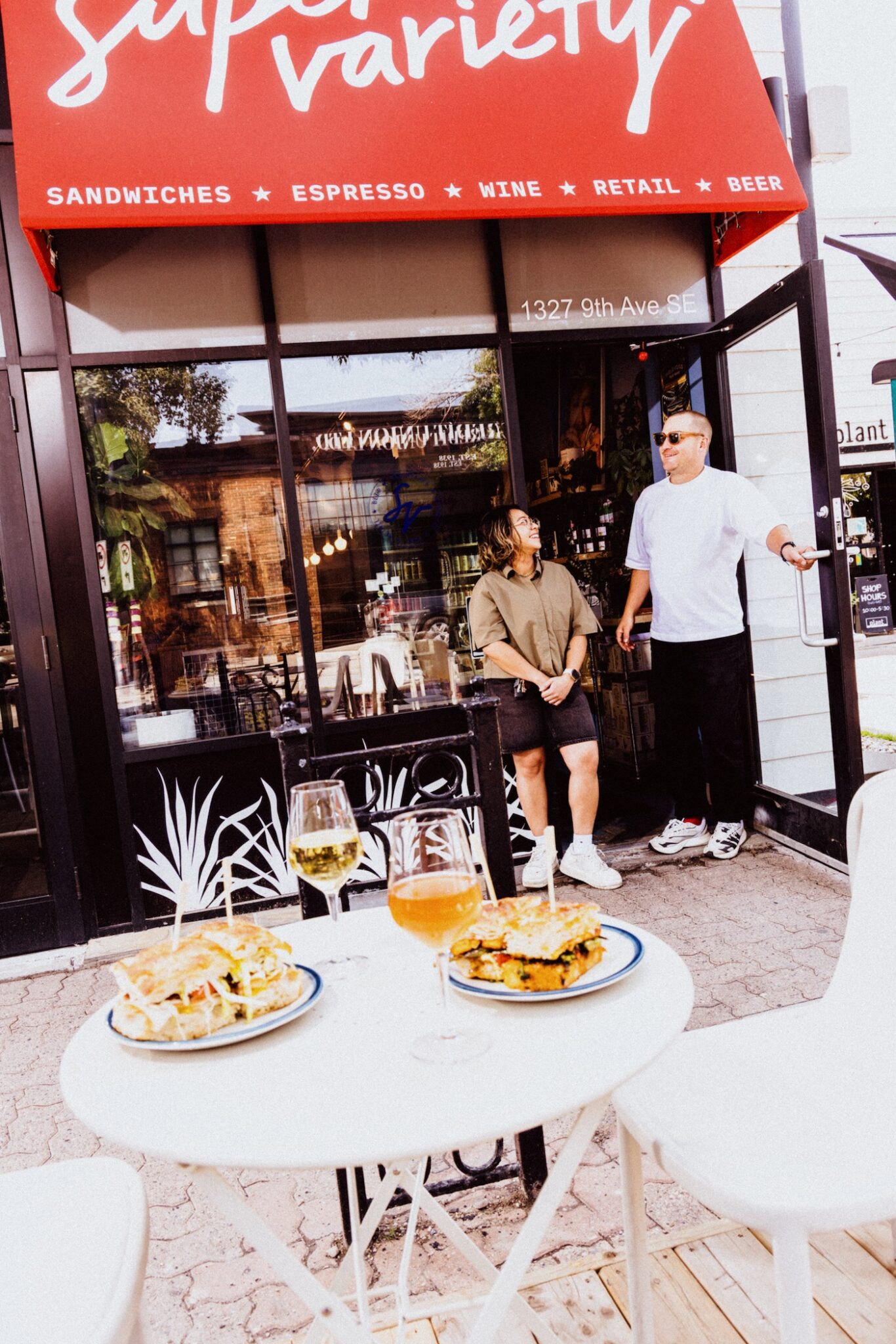 Two people stand outside the door to Super Variety near an outdoor table set up with two sandwiches and drinks.