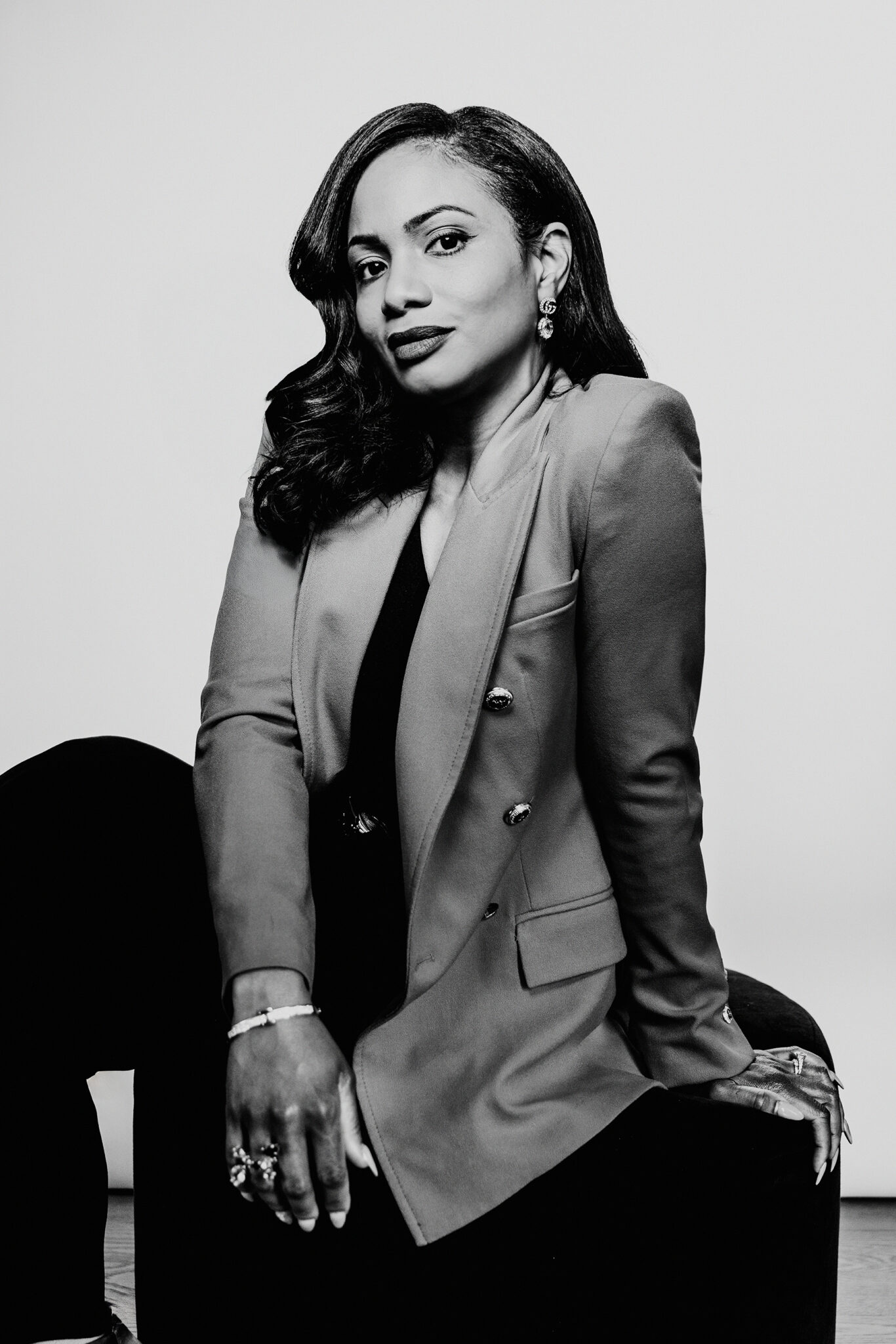 A black and white portrait of a woman wearing a blazer sitting on a stool.