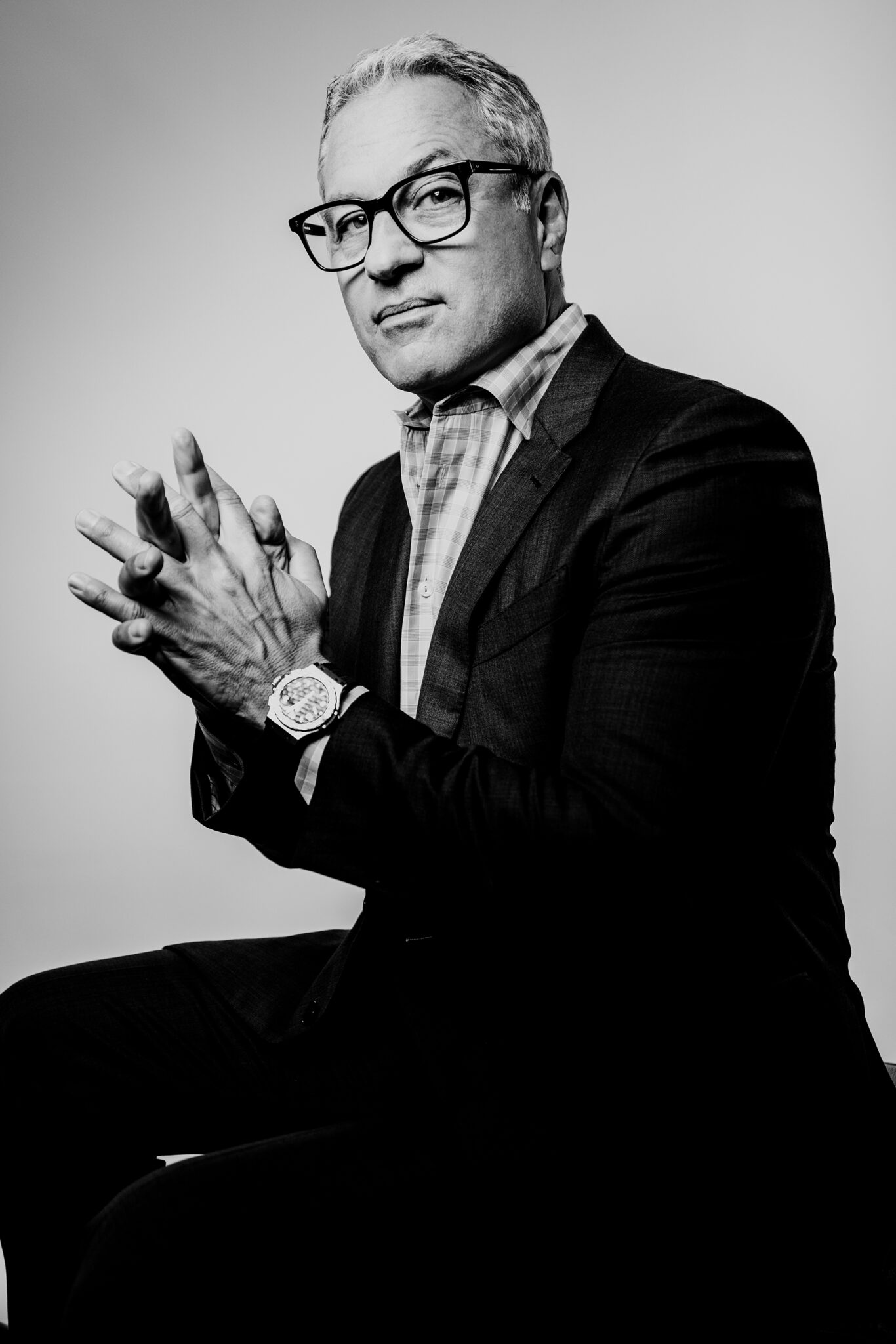 A black and white portrait of a man wearing a suit and glasses with his fingers crossed.