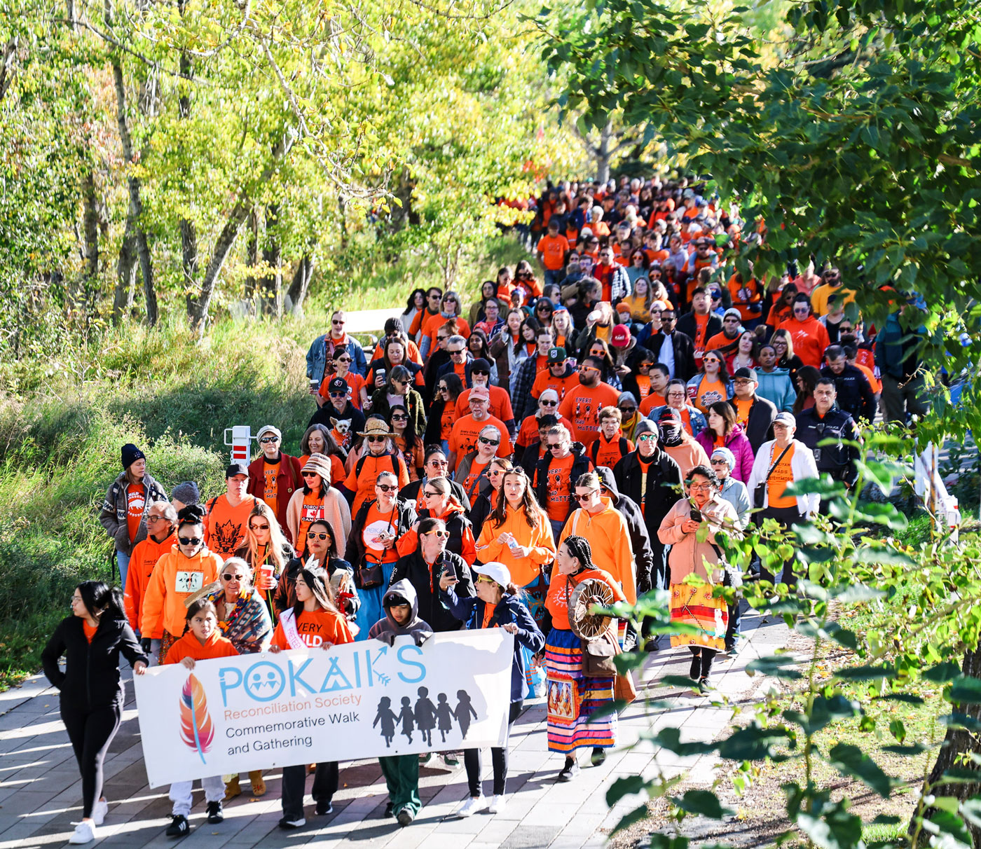 A large group wearing orange shirts walks through a green area for the Pokaiks Reconciliation Society's Commemorative Walk and Gathering