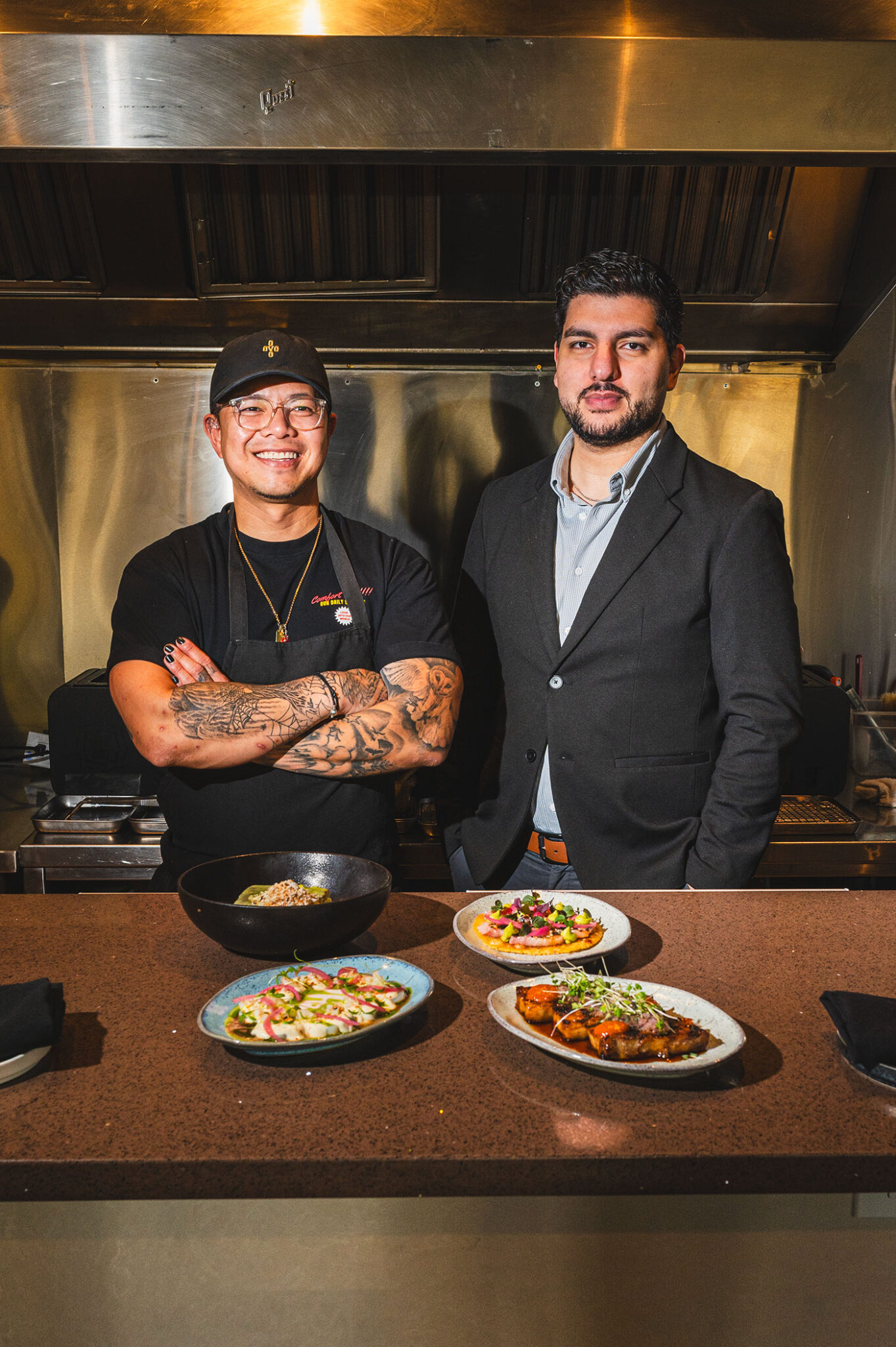 Two men stand inside a small kitchen with dishes of food on the table in front of them.