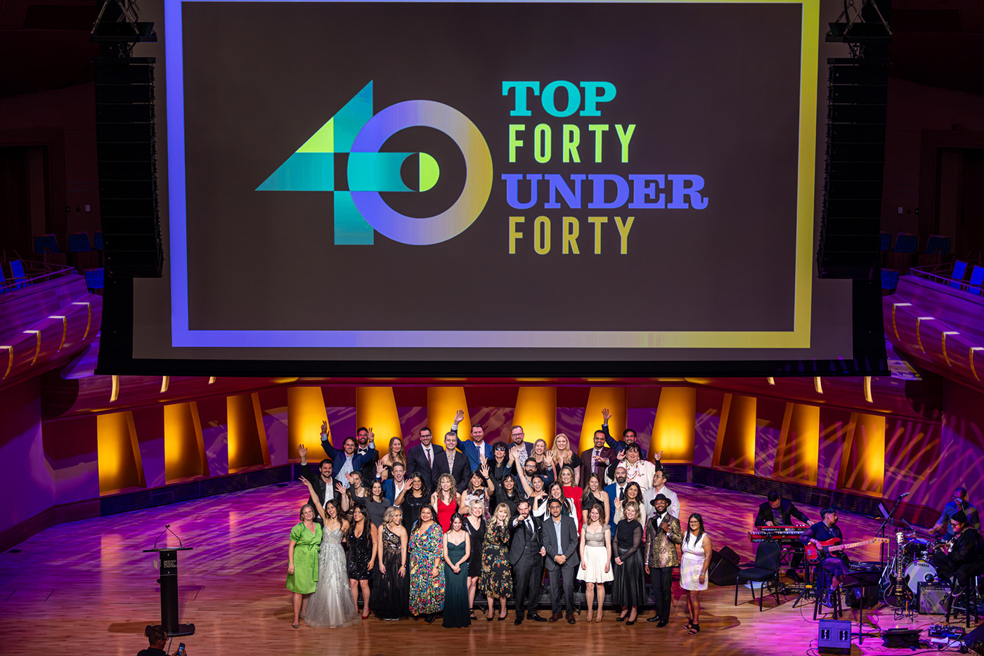 A group of people stand together on a stage under a screen saying Top 40 Under 40