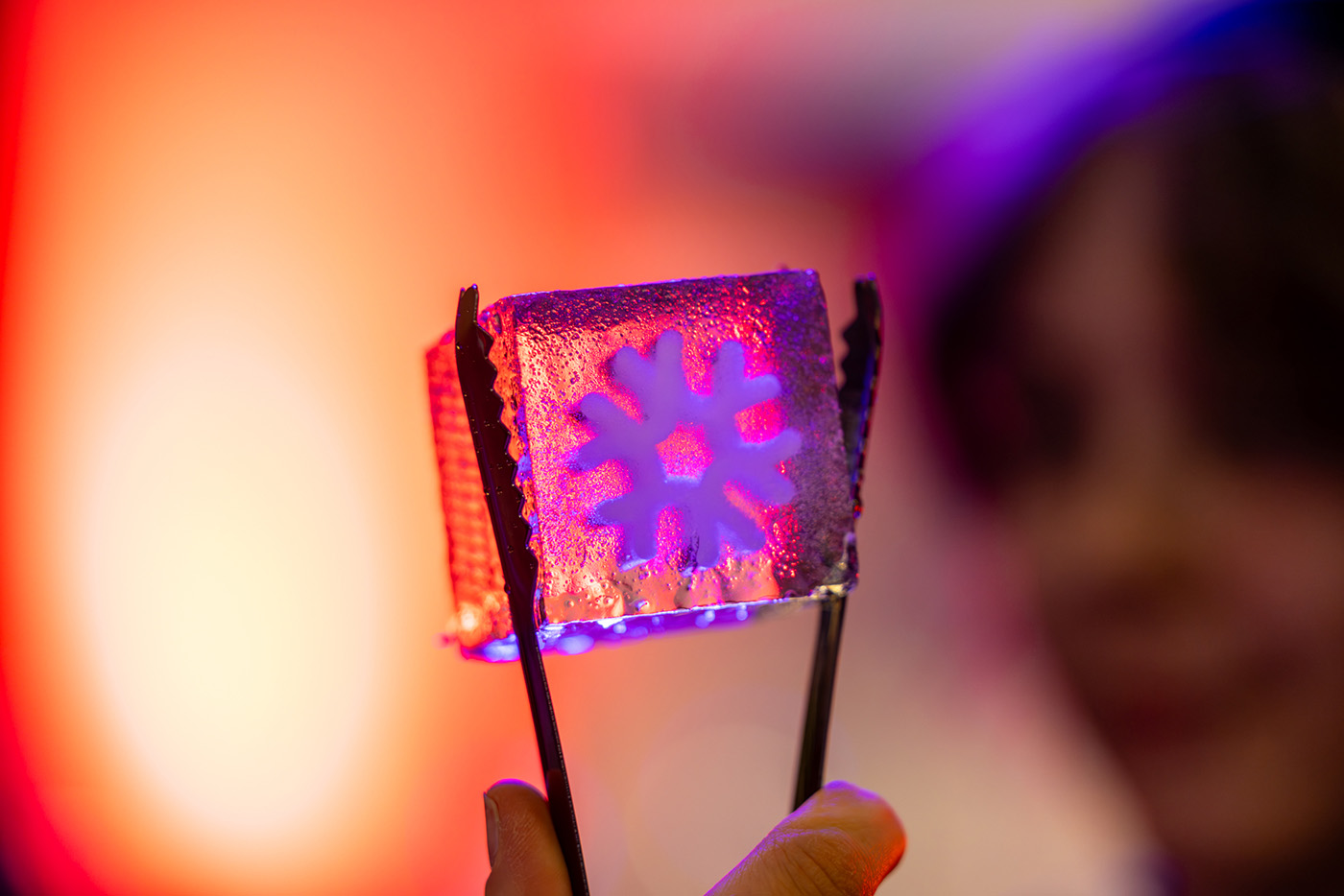 Tongs hold up an ice cube with a snowflake design in the middle.